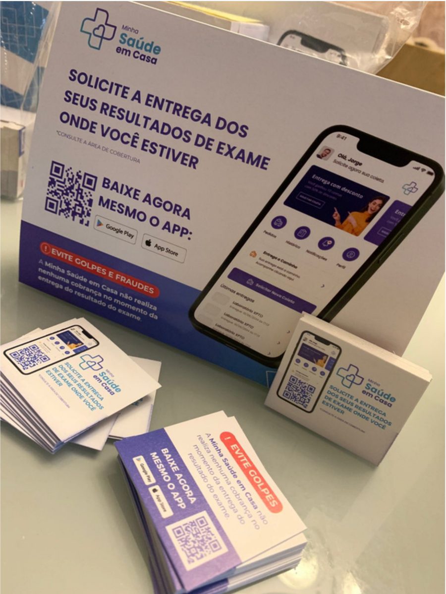 Display de mesa e cartões da Minha Saúde em Casa na recepção de um laboratório, com QR code para baixar o app